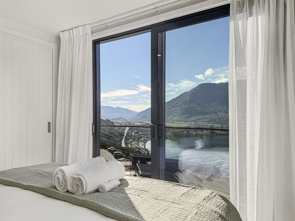 A hotel room with a bed, a view of a mountainous landscape through a large window, and a balcony with a view of a river and mountains.