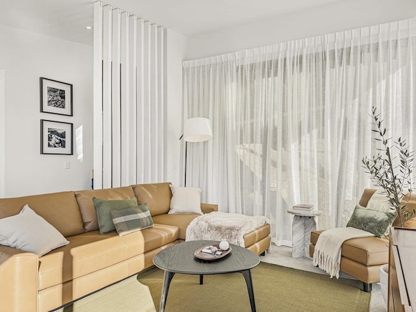 A modern living room with a sofa, a coffee table, and a side table. There are curtains and a lamp.