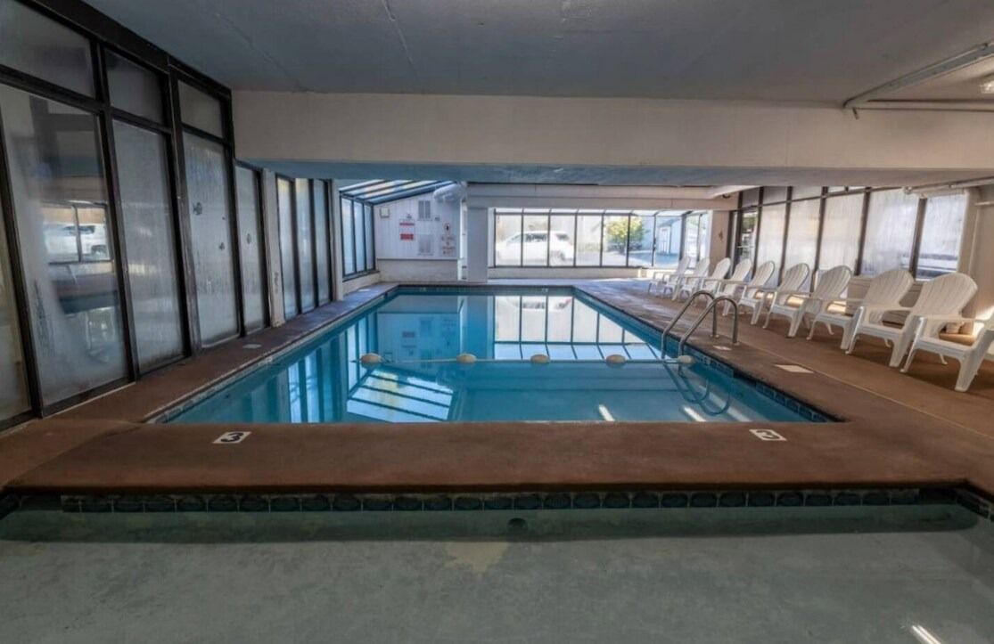 Une piscine dont le bord est revêtu de carrelage, entourée de chaises longues blanches, et offrant une vue sur l’extérieur grâce à de grandes fenêtres.