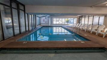 Une piscine dont le bord est revêtu de carrelage, entourée de chaises longues blanches, et offrant une vue sur l’extérieur grâce à de grandes fenêtres.