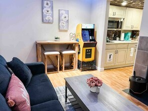 Een woonkamer met een bank, een salontafel met bloemen, een eethoek met stoelen, een keuken met kasten, en een spelconsole.