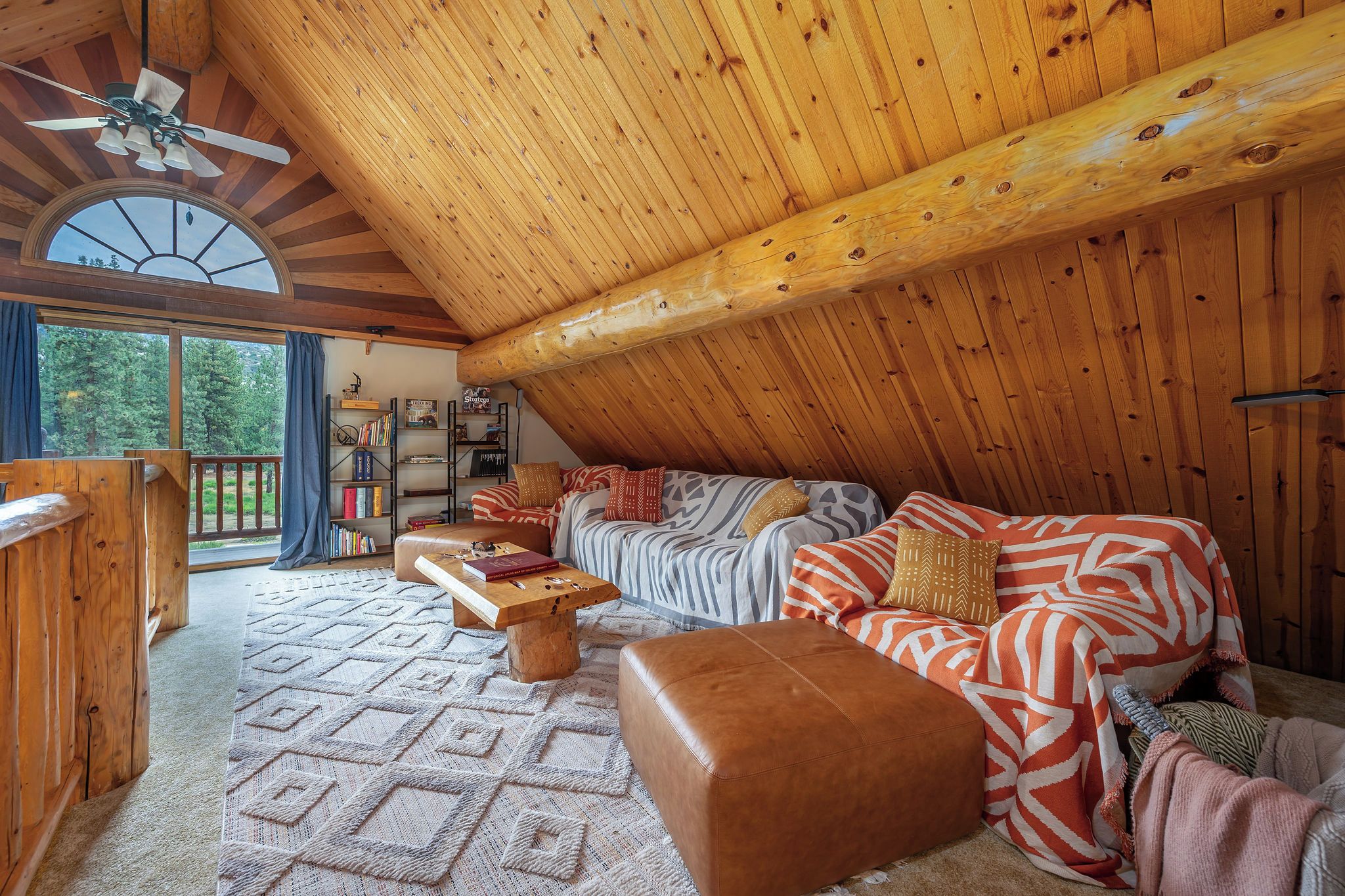 Una habitación estilo loft acogedora, con techo inclinado, vigas de madera, un ventanal grande y una estantería empotrada.