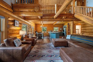 Interior de una cabaña de troncos con sala de estar, comedor, cocina y una escalera.