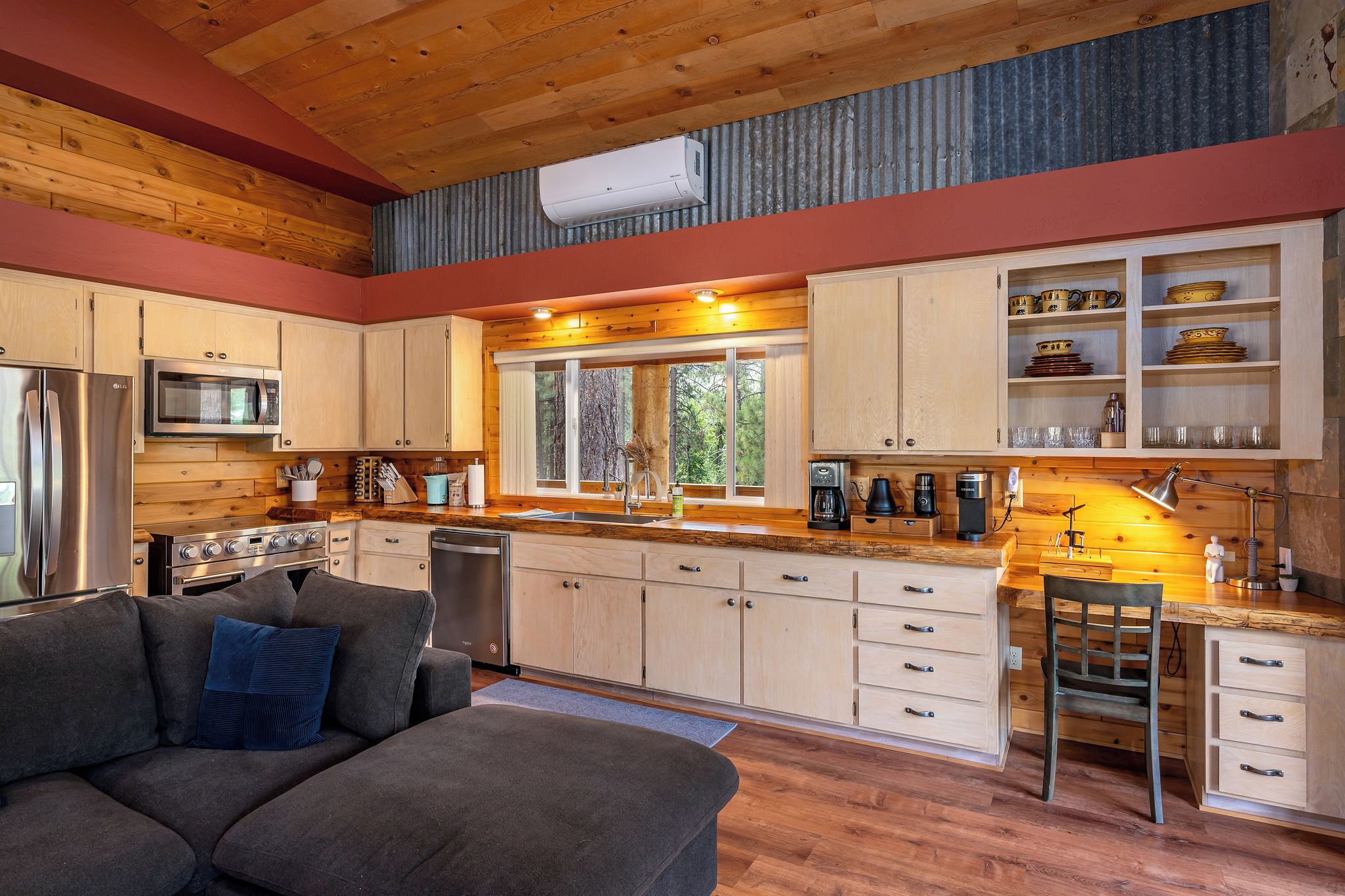 A modern kitchen with wooden cabinets, stainless steel appliances, and a large window overlooking a forest.