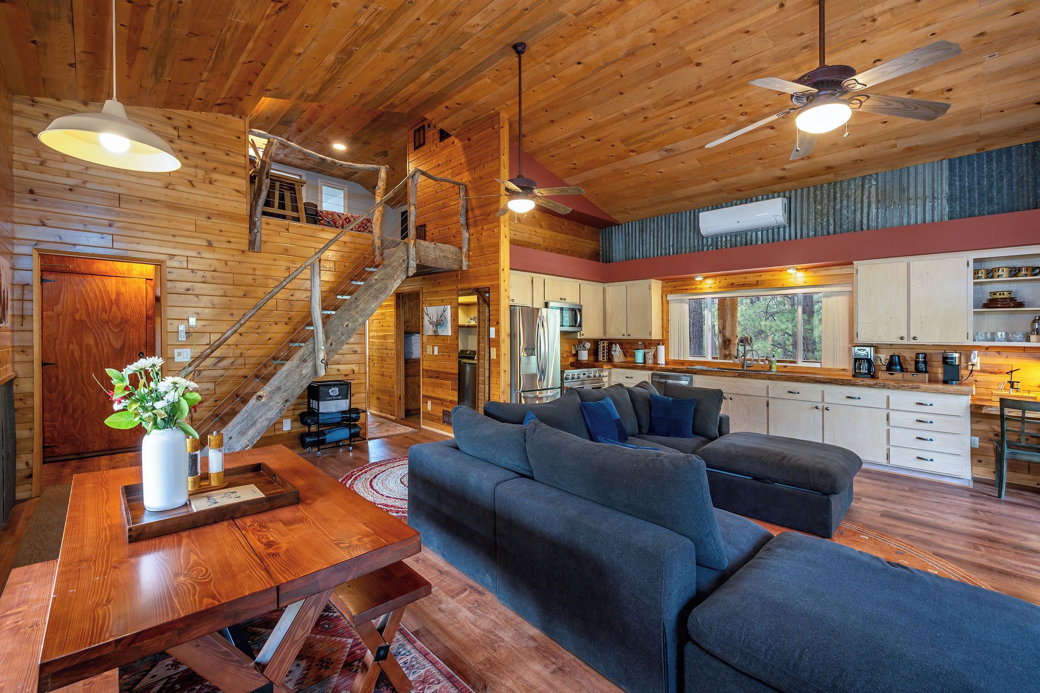 A spacious living area with a wooden staircase, a sofa set, a dining table, and a kitchen area.
