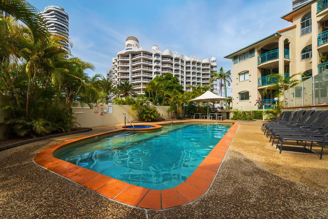 A swimming pool with a pebble border, surrounded by lounge chairs and palm trees, with a hotel building in the background.