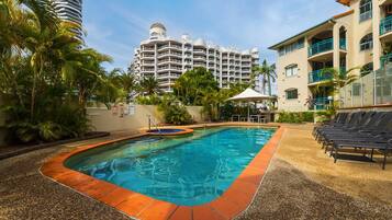 A swimming pool with a pebble border, surrounded by lounge chairs and palm trees, with a hotel building in the background.