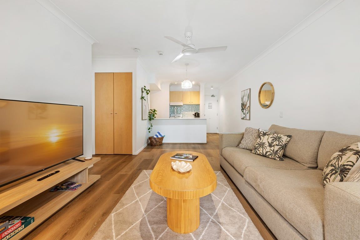 A living room with a television, sofa, coffee table, and a kitchen in the background.