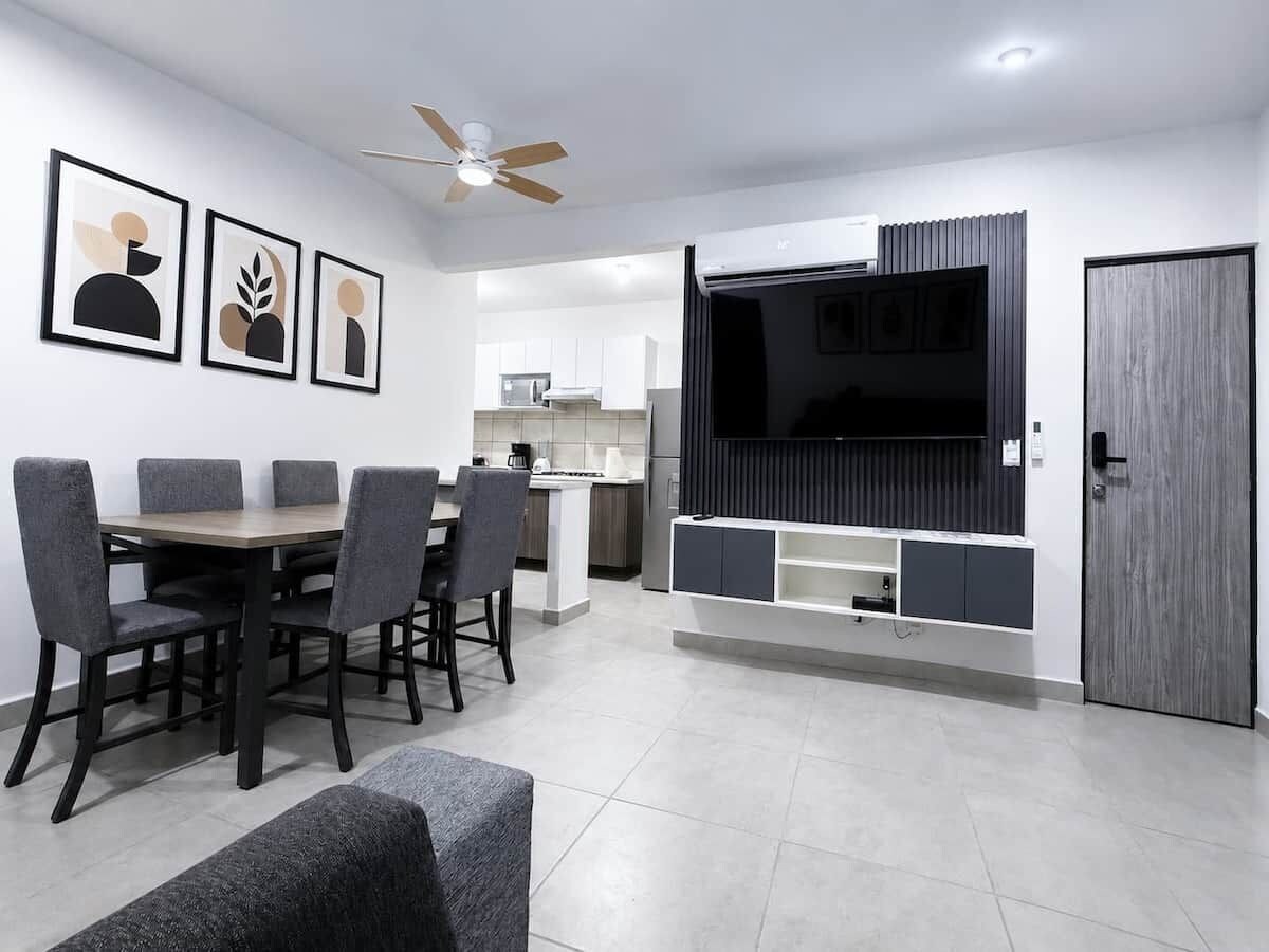 A modern living room with a dining area, a kitchen in the background, and a large flat-screen TV mounted on a wall.