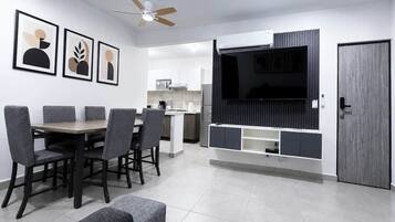 A modern living room with a dining area, a kitchen in the background, and a large flat-screen TV mounted on a wall.