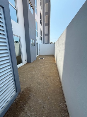 Un estrecho pasillo exterior con suelo de tierra, bordeado por una pared blanca y un edificio con varias ventanas.