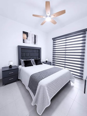 Une chambre à coucher avec un lit, des tables de chevet, un ventilateur de plafond et une fenêtre avec des stores.