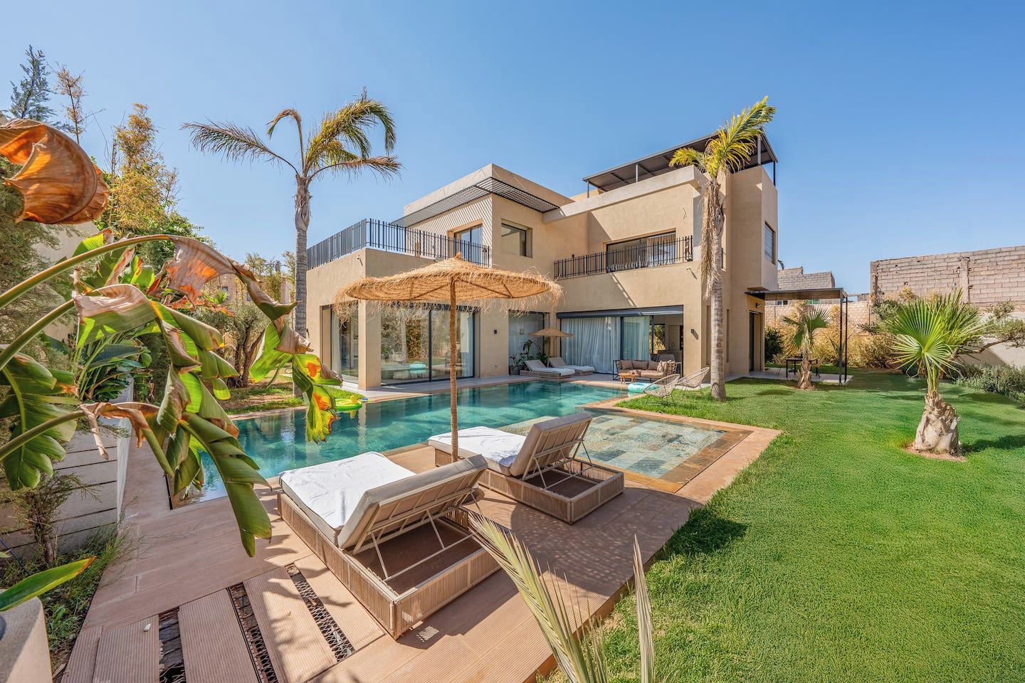 Una casa moderna de dos pisos con piscina, mobiliario exterior y plantas tropicales.
