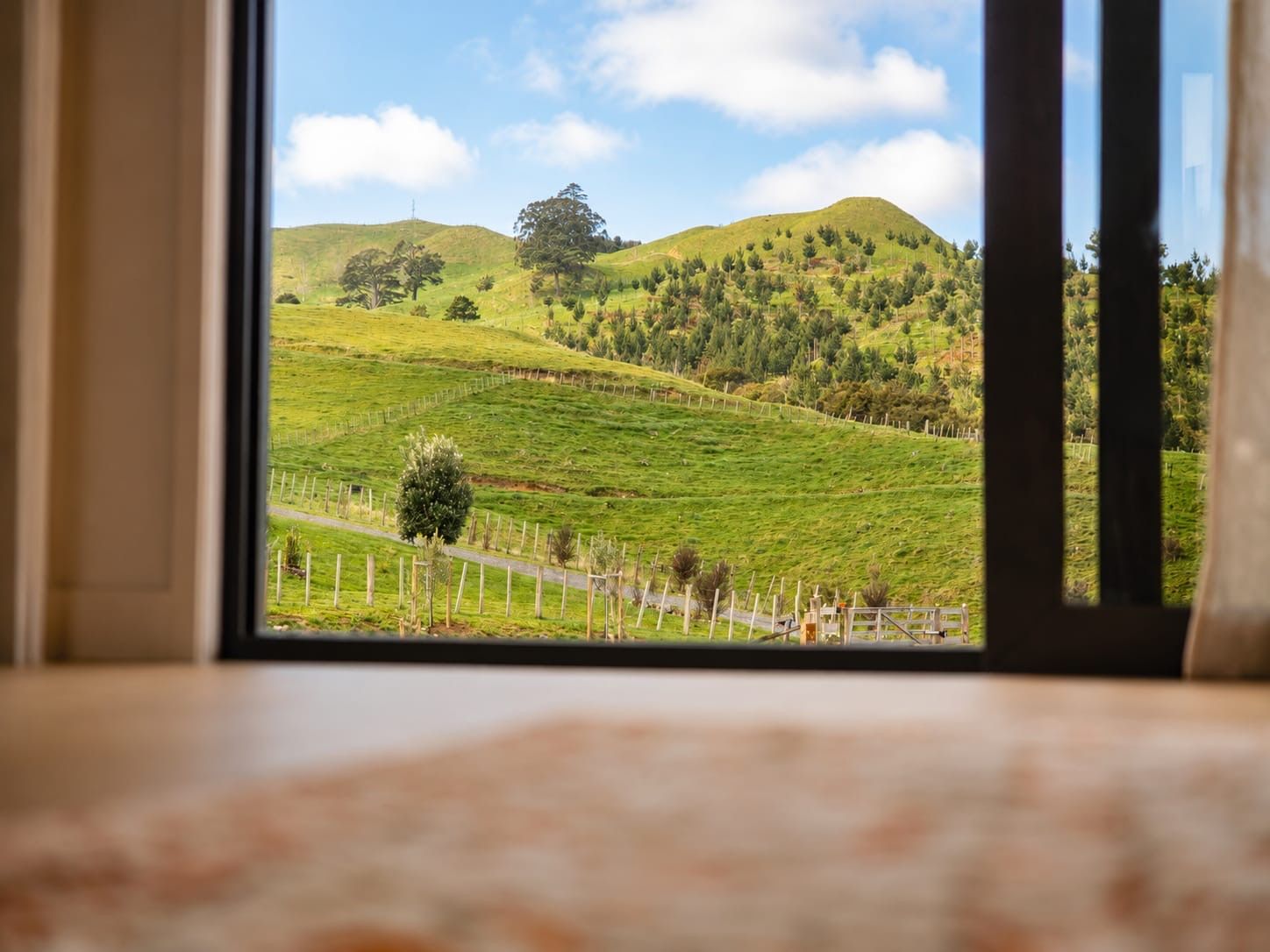 A view through a window of a green landscape with a fence, trees, and a hill.