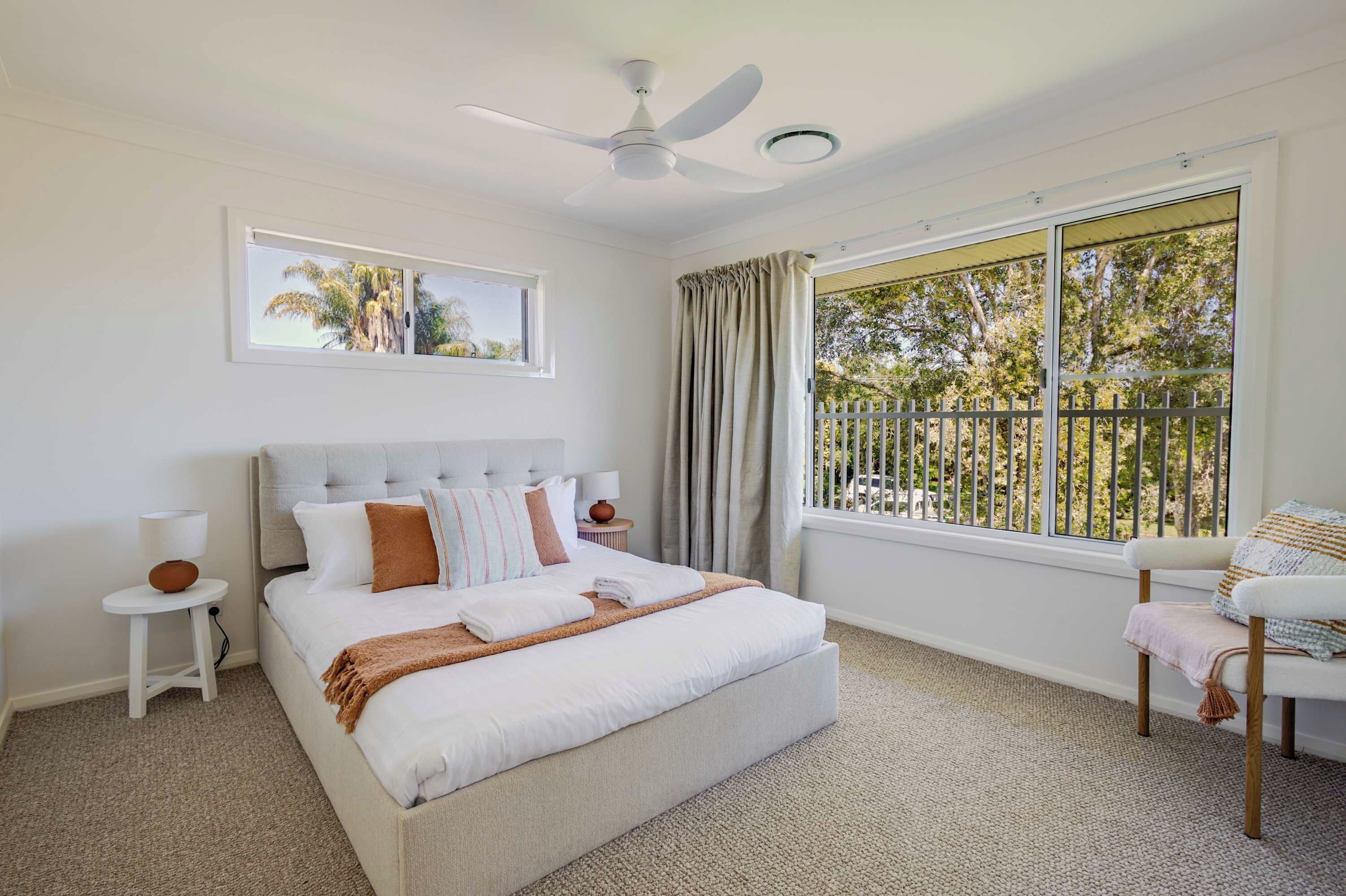 A bedroom with a bed, a ceiling fan, a window with curtains, a chair, and a small table with a lamp.