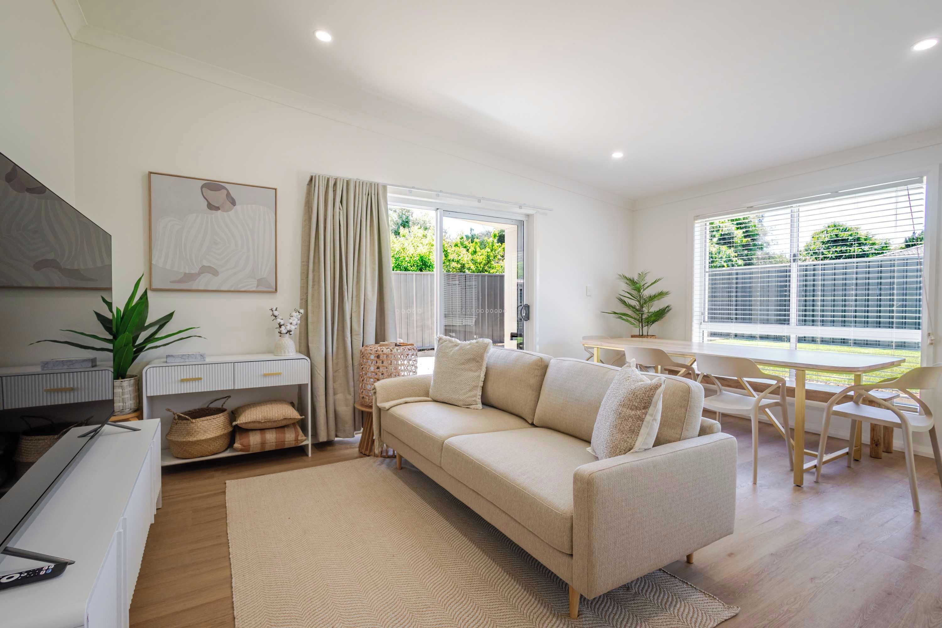 A modern living room with a sofa, a TV, a dining area with chairs, and a large window with a view of greenery.
