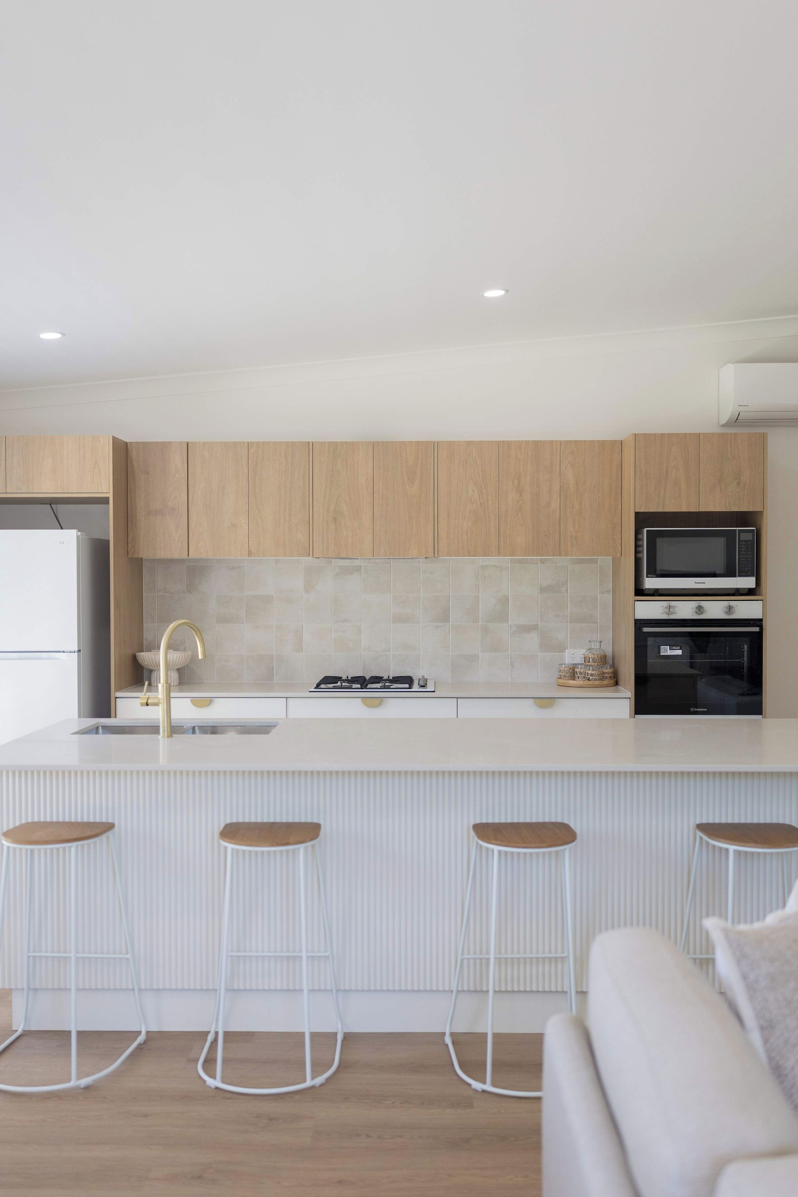Una cocina moderna con armarios de madera, encimera blanca, estufa a gas, pileta y microondas integrado. Hay tres taburetes blancos con asientos de madera.