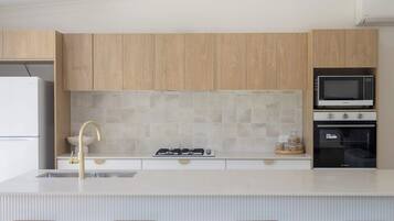 Una cocina moderna con armarios de madera, encimera blanca, estufa a gas, pileta y microondas integrado. Hay tres taburetes blancos con asientos de madera.