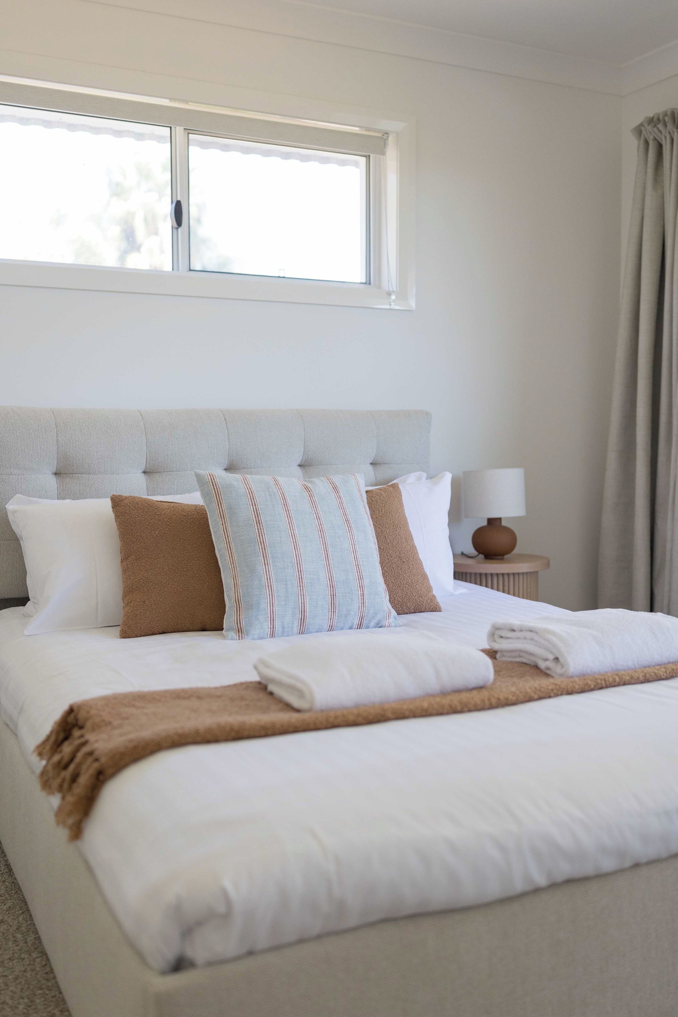 A neatly made bed with pillows and a blanket, a bedside table with a lamp, and a window with curtains.