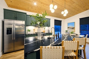 A modern kitchen with a central island, stainless steel appliances, and a dining area with white chairs. - Shake It Off · Couple`s Family Cabin: Hot Tub, Playground, & Fire-Pit (Broken Bow)