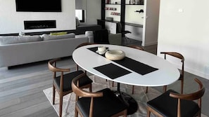 A modern living room with a white sofa, a flat-screen TV mounted on the wall, and a dining area with a white table and black chairs. - Daylesford Scandinavian Chalet (Daylesford)
