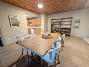 A dining area with a wooden table and chairs, a kitchen in the background, and a shelving unit with various items. - Blacksburg Cranberry Lodge - VT Luxury Retreat (Blacksburg)