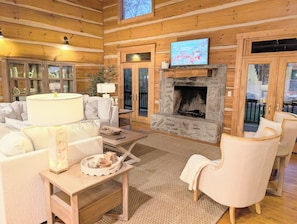 Interior acolhedor de uma cabana com lareira de pedra, paredes de madeira, televisão e uma área de estar confortável.