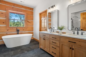 A modern bathroom with a freestanding bathtub, wooden cabinetry, a large mirror, and a window with a view of trees. - Blacksburg Cranberry Lodge - VT Luxury Retreat (Blacksburg)