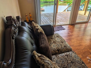 A dark leather sofa with patterned cushions, a small table with figurines, and a view of a wooden deck and water outside. - Lakefront 3-bedroom house with AC in cozy Eatonton (Eatonton)