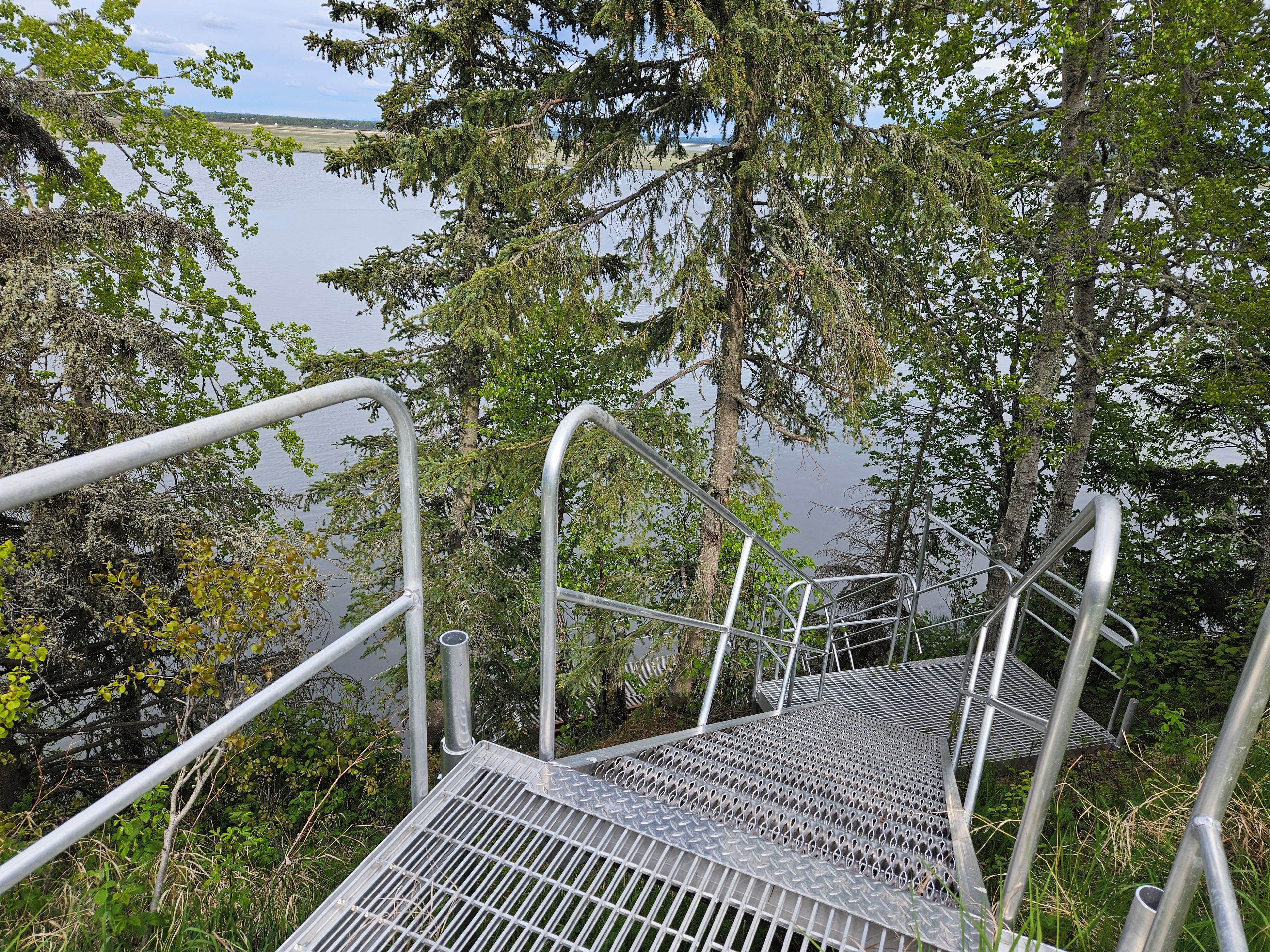 Freiliegende Metalltreppen führen zu einer Aussichtsplattform mit Metallgeländern, umgeben von Bäumen und mit Blick auf ein Gewässer.