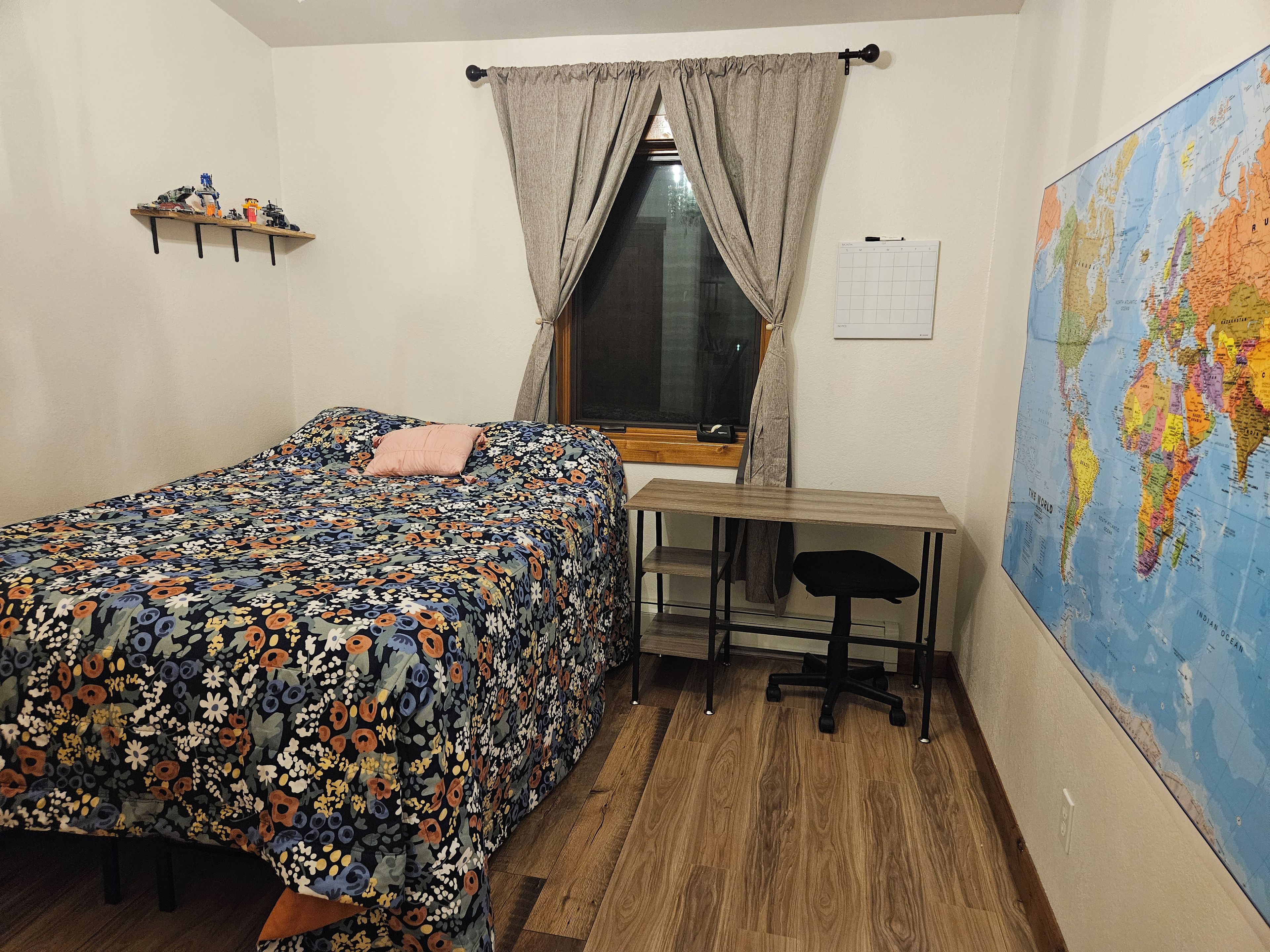Un dormitorio con una cama, un escritorio, una silla, un ventanal, un mapa en la pared y un estante con objetos.