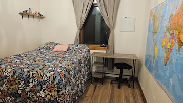 Un dormitorio con una cama, un escritorio, una silla, un ventanal, un mapa en la pared y un estante con objetos.