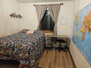 Un dormitorio con una cama, un escritorio, una silla, un ventanal, un mapa en la pared y un estante con objetos.