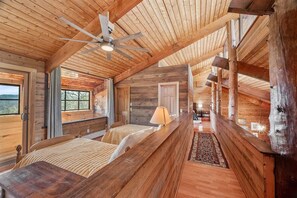 Interior de uma cabana de madeira com uma cama central, um ventilador de teto e vista pelas janelas.