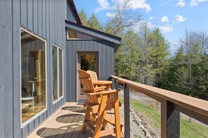 Um deck de madeira com cadeiras Adirondack, porta de vidro e vista para árvores e céu.