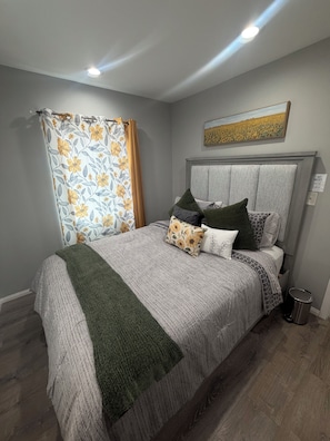 A bedroom with a bed, a headboard, a painting, a curtain, and a trash bin. -  2-bedroom apartment with gated parking (Los Angeles)
