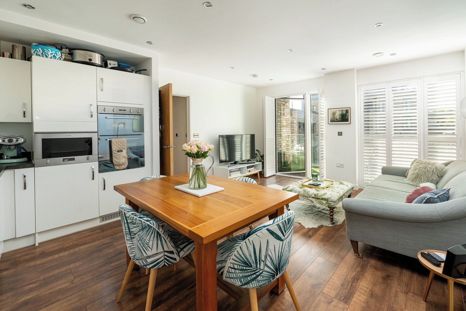 A modern kitchen with built-in appliances, a dining area with a wooden table and patterned chairs, a sofa, and a TV in a living area with a sliding glass door to a balcony.
