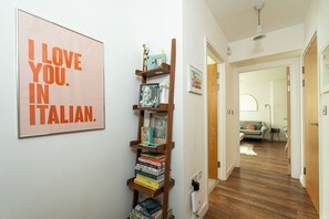 A hallway with a bookshelf, a framed poster, and a glimpse of a living room with a sofa. - Bright 1BR Flat with Balcony in London Fields (London)
