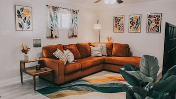 Uma sala de estar com um sofá seccional, uma mesinha lateral, um abajur e uma planta grande.