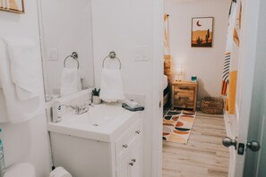 Casa de banho com lavatório branco, dois espelhos, porta-toalhas e uma pequena mesa de cabeceira com uma lâmpada.