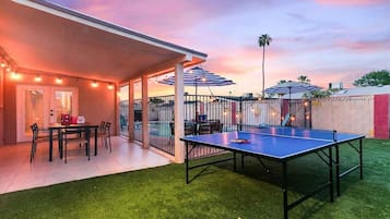 Un patio exterior con mesa de ping pong, zona de estar techada y luces colgantes.