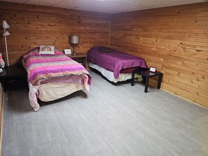 Ein Zimmer mit zwei Betten, Holzwänden, einem Nachttisch, einer Lampe und einem kleinen Tisch.