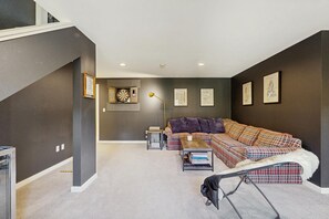 Ein modernes Wohnzimmer mit einer Eckcouch, einem Couchtisch und einer Lampe. An den Wänden hängen gerahmte Bilder und es befindet sich eine Dartscheibe im Raum.