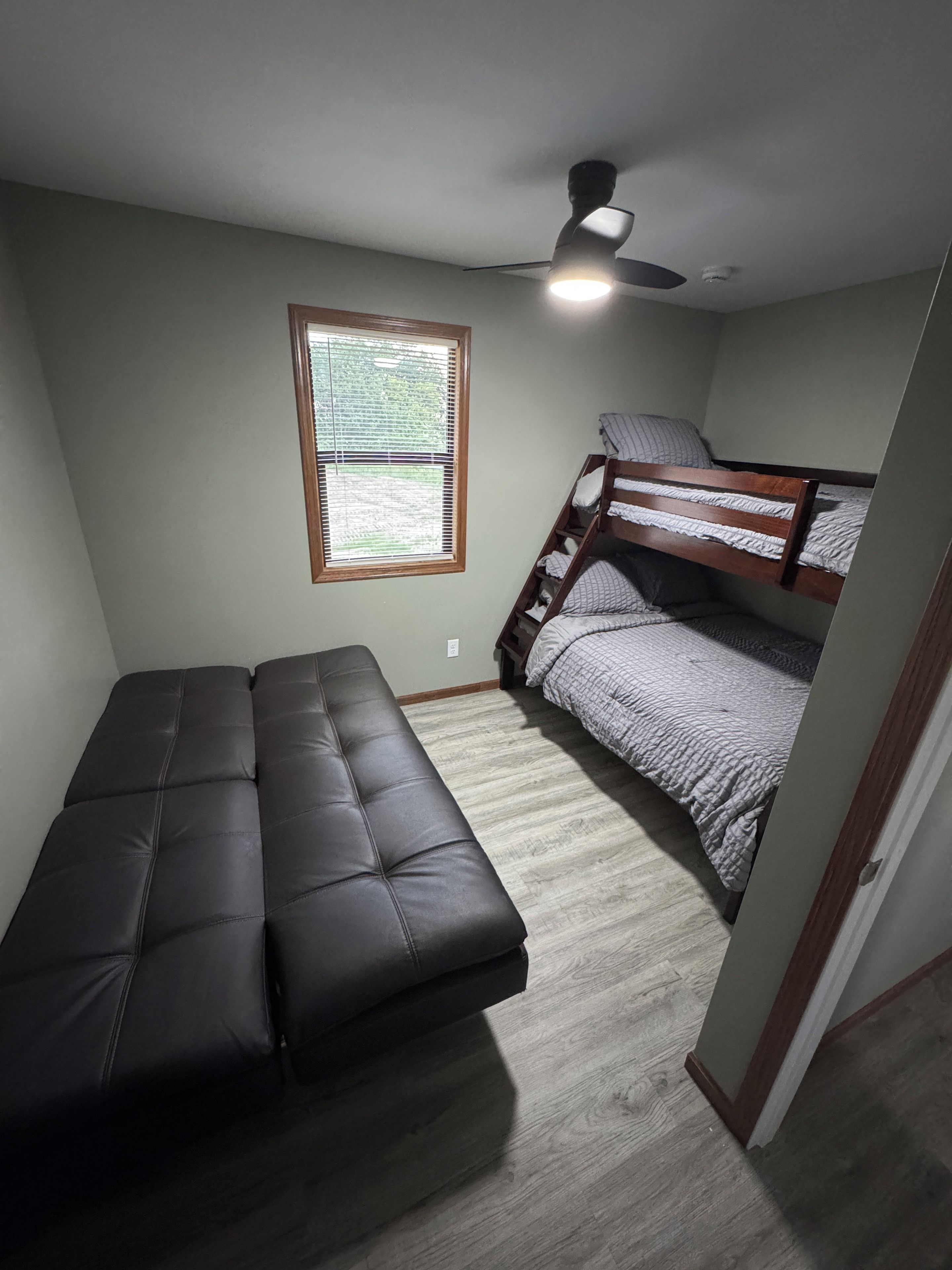 A room with a bunk bed, a sofa, a ceiling fan, and a window with blinds.