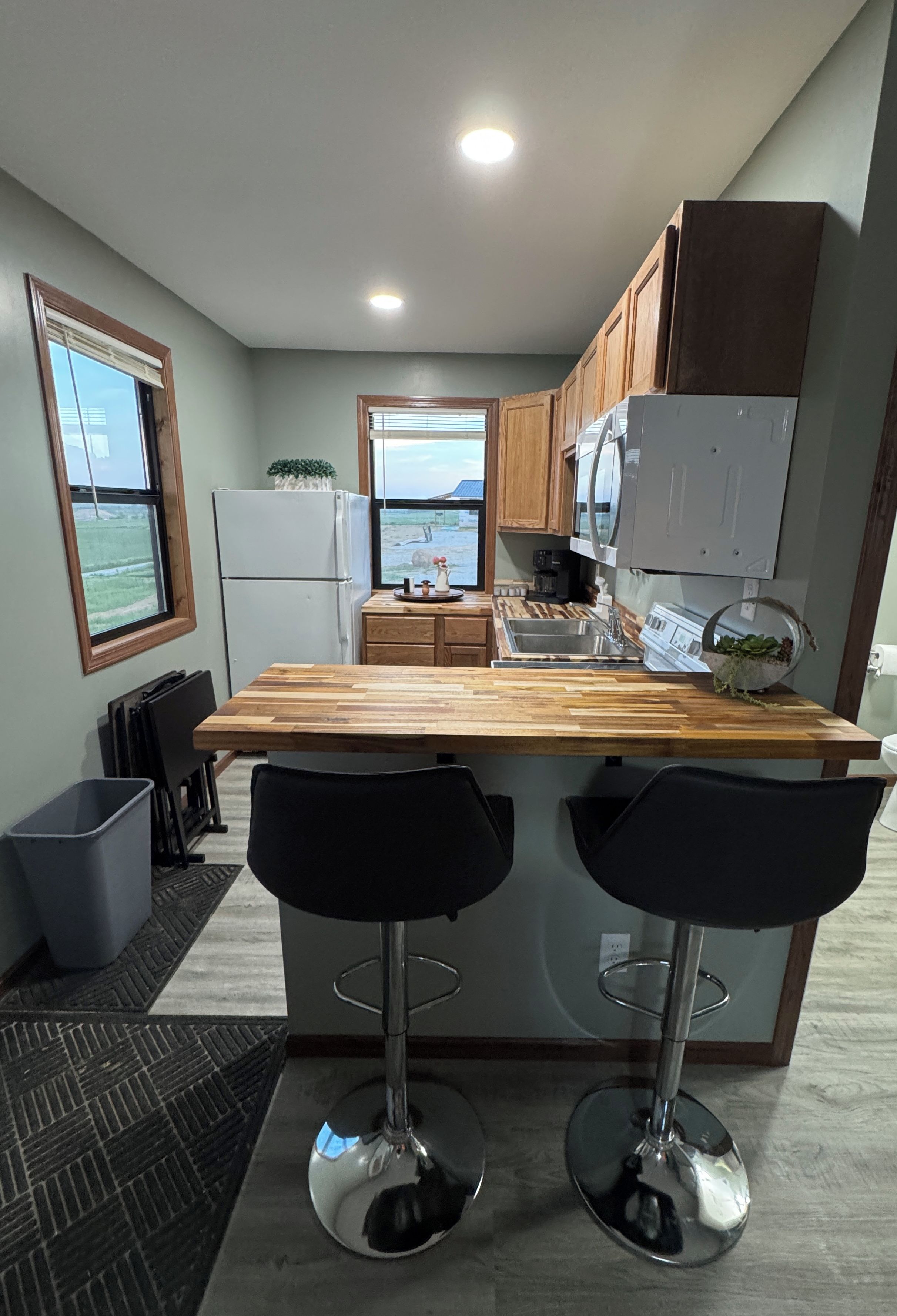 A modern kitchen with a wooden island, two bar stools, a refrigerator, and a view of the outdoors.