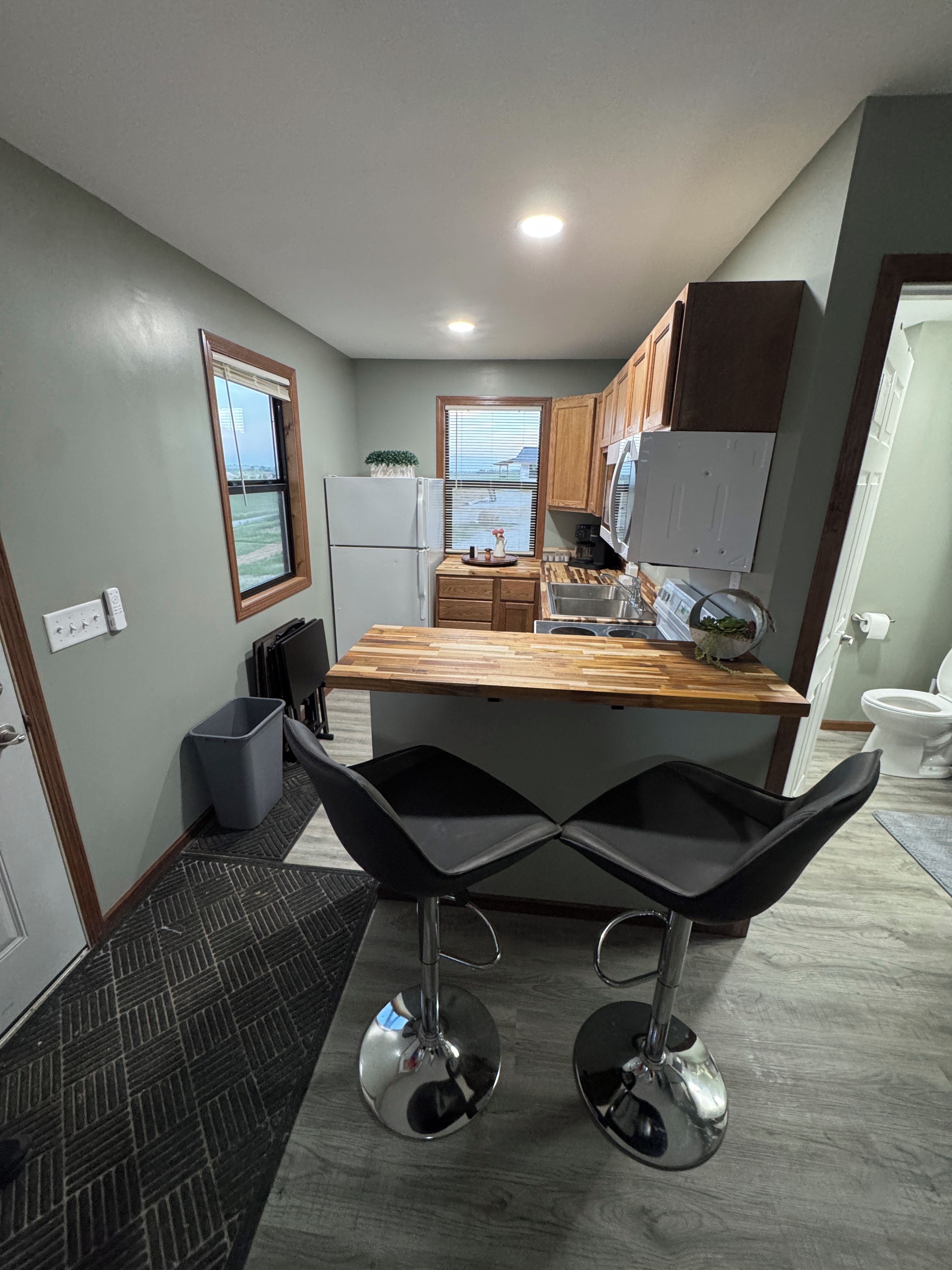 A modern kitchen with a central island, bar stools, wooden cabinets, and a view through the window.