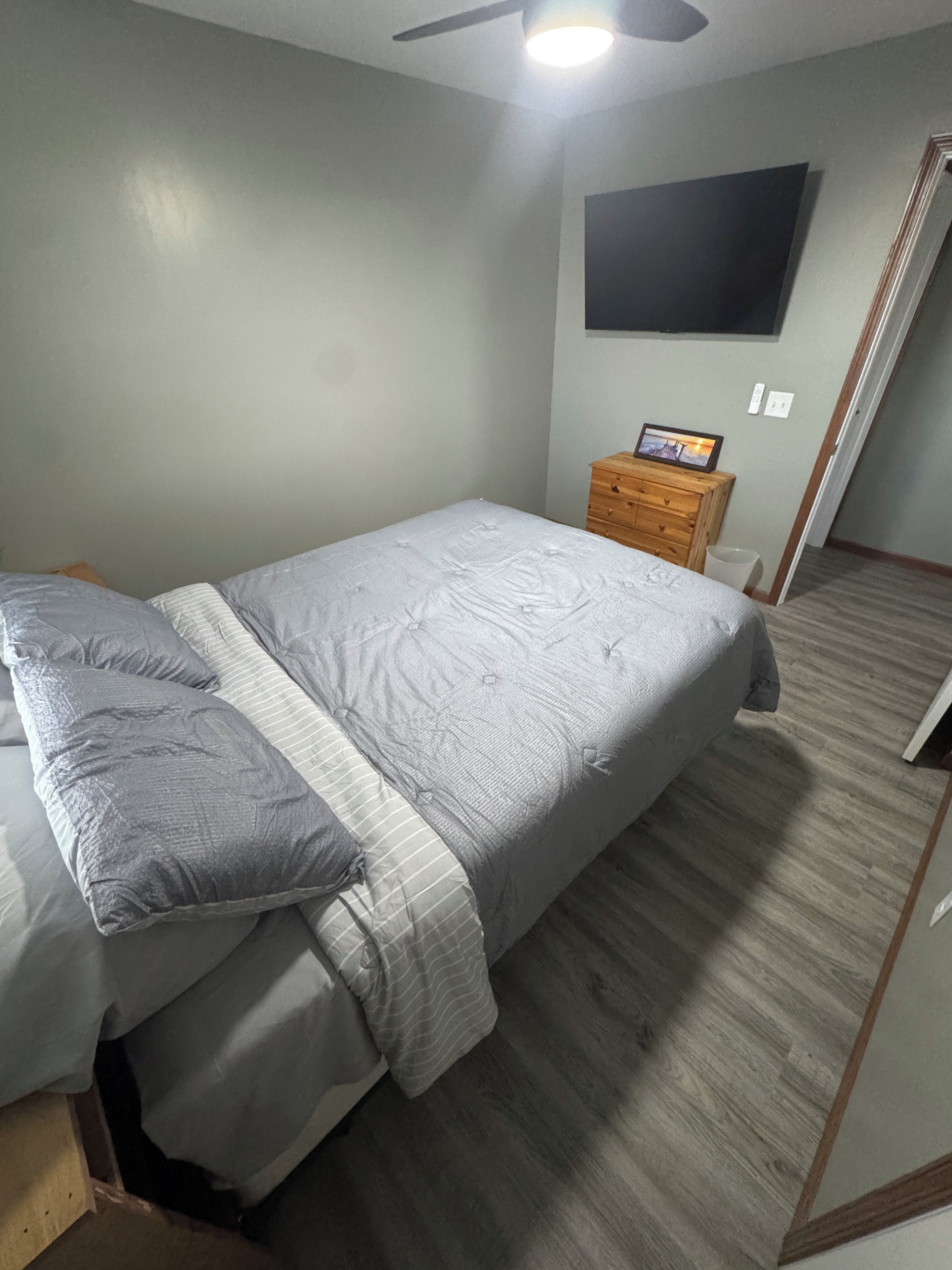 Une chambre à coucher avec un lit, une table de chevet en bois, un tableau encadré et un ventilateur de plafond.