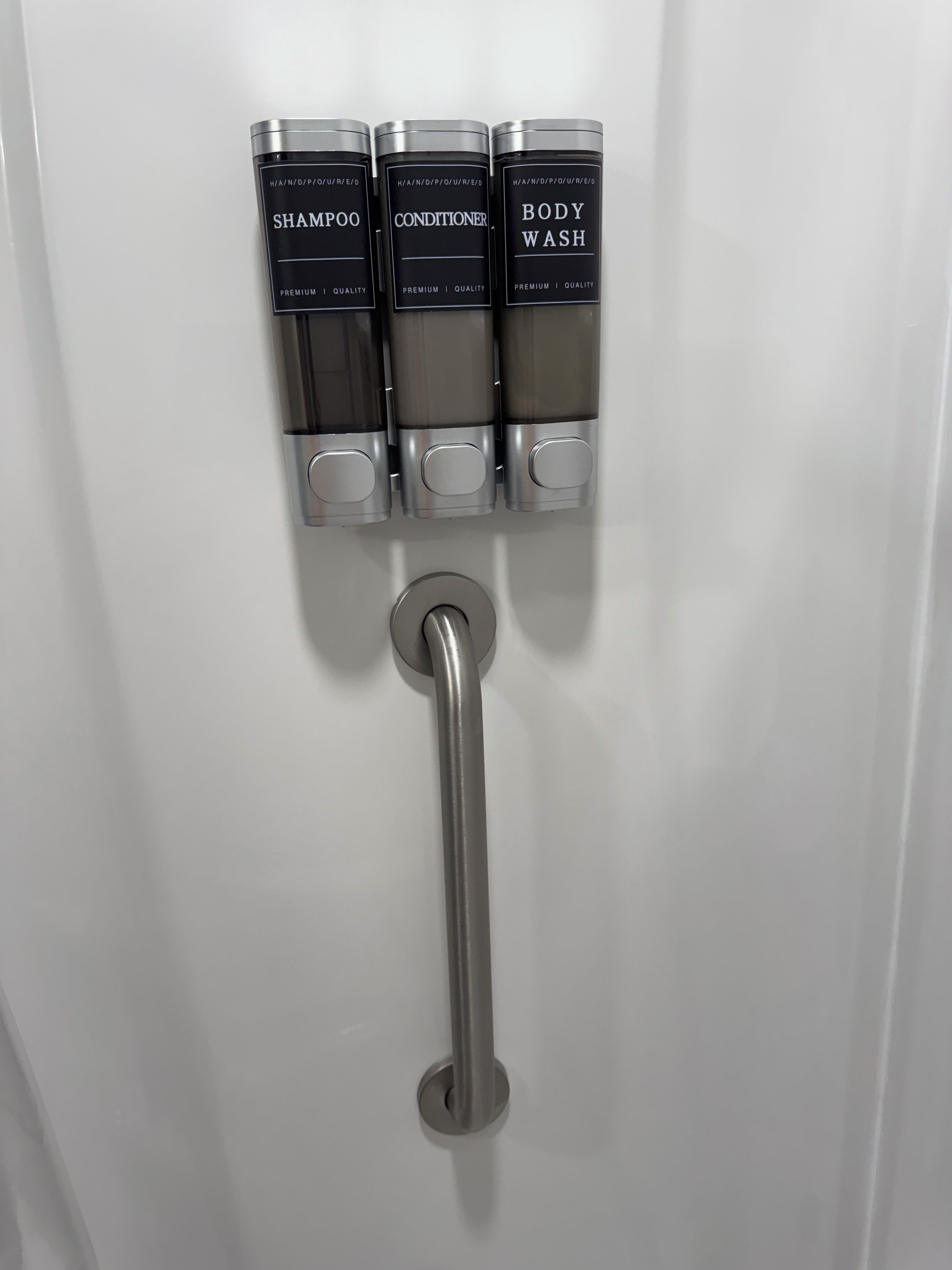 Eine wandmontierte Spenderstation mit drei Fächern, beschriftet mit 'SHAMPOO', 'CONDITIONER' und 'BODY WASH'.