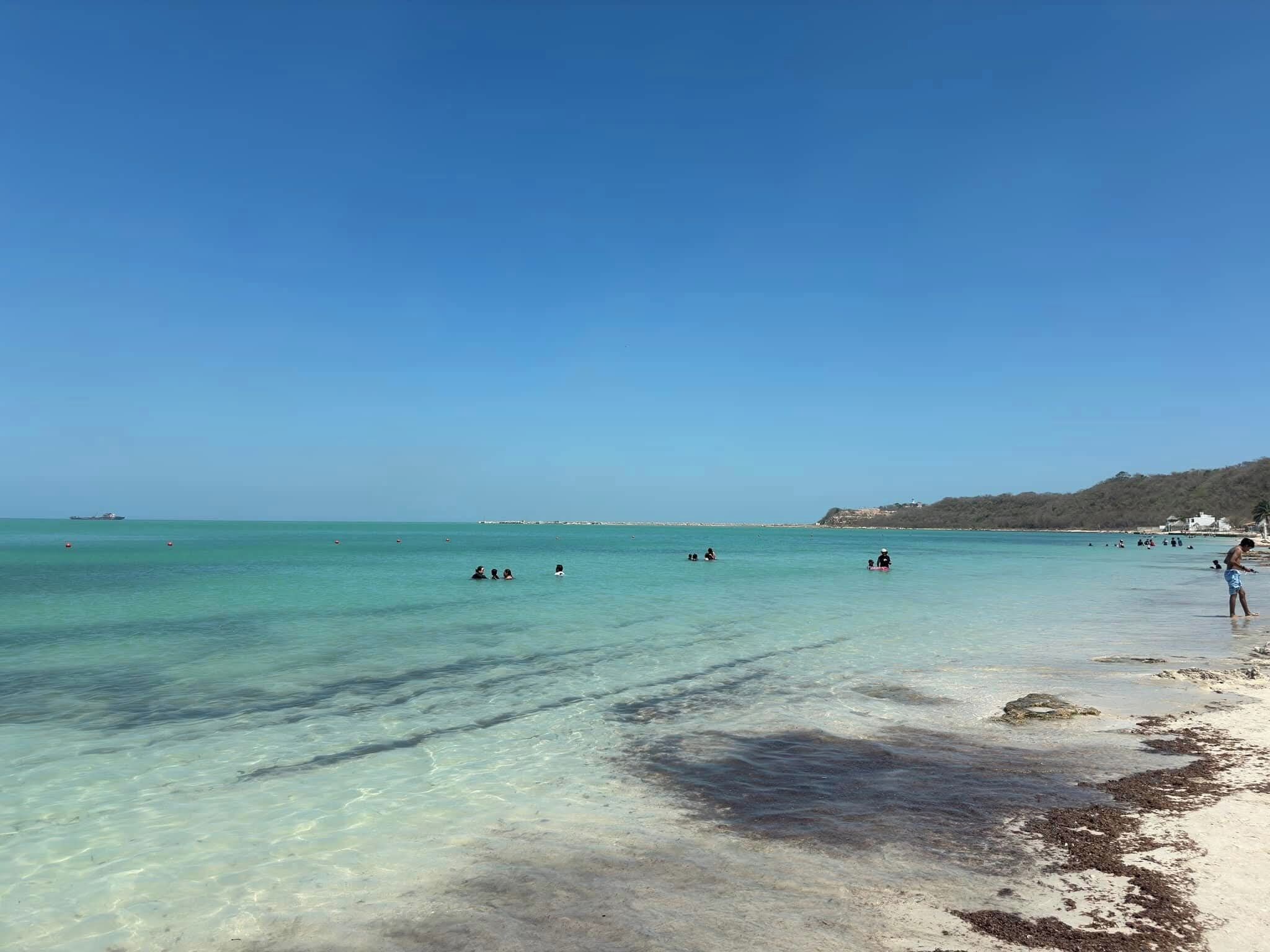 A beach with clear turquoise water, a sandy shore, and a few people swimming.