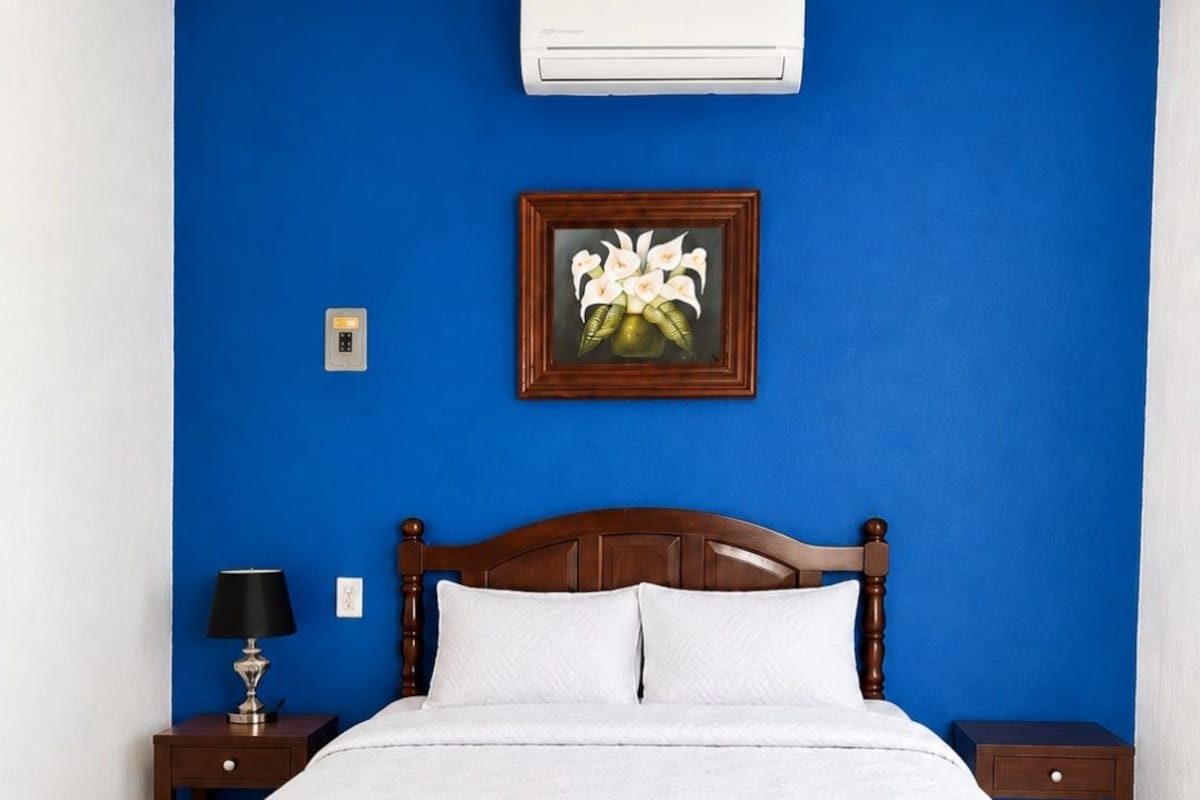 A bedroom with a bed, a bench, a nightstand, a lamp, and an air conditioner.